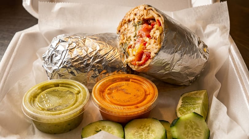 Two foil-wrapped burritos with salsa cups, cucumber slices, and lime in a takeout container.