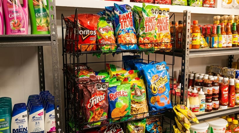 A store shelf displays various snack bags, hot sauces, and personal care products.
