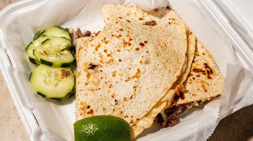 Three quesadillas with meat and cheese, sliced cucumbers, and a lime wedge in a takeout container.