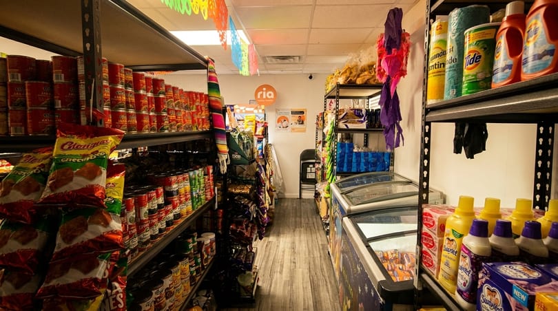 A narrow grocery store aisle with shelves stocked with canned goods, snacks, and household items.